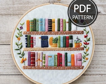 May include: Embroidered hoop art featuring a bookshelf with colorful books, green plants, and a small cup. The hoop is white with a wooden frame. The text "PDF PATTERN" is in a black circle on the top right corner of the image.