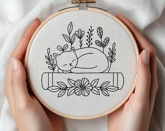 Puede incluir: Un bastidor de bordado con un dibujo lineal negro de un gato durmiendo sobre un libro, rodeado de elementos florales. El diseño está sobre una tela color crema, sujeta en un bastidor de madera. El diseño es simple y encantador.