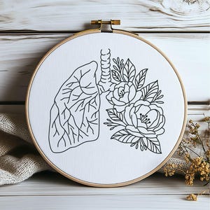 Peut inclure: Un cercle à broder noir et blanc représentant des poumons et des fleurs. Le motif est sur un tissu blanc, maintenu dans un cercle en bois. L'œuvre combine des éléments anatomiques et botaniques, pour la décoration.