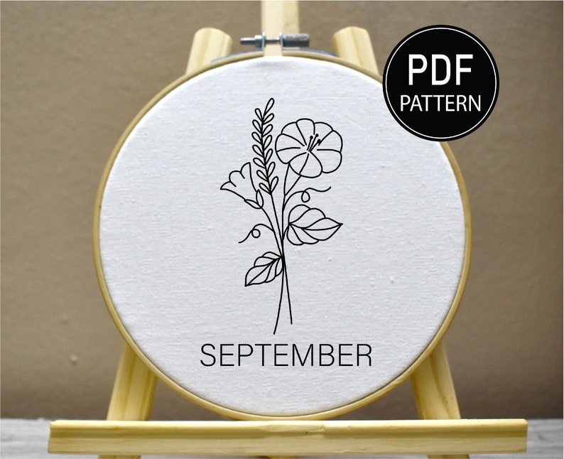 Puede incluir: Un aro de bordado blanco con un dibujo de l&iacute;nea negra de una flor y hojas. El aro est&aacute; en un caballete de madera y el texto "SEPTEMBER" est&aacute; debajo del dibujo. El texto "PDF PATTERN" est&aacute; en un c&iacute;rculo negro en la esquina superior derecha de la imagen.