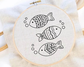 May include: Three black embroidered fish designs on a white fabric background in a wooden embroidery hoop.