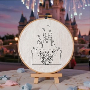 Puede incluir: Un bastidor de bordado redondo con un dibujo lineal negro de un castillo con orejas de Mickey Mouse y detalles florales. El bastidor se exhibe en un pequeño caballete de madera, con un fondo borroso de un castillo y fuegos artificiales.