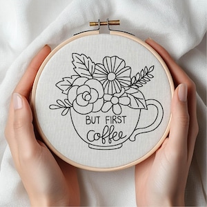 May include: An embroidery hoop featuring a floral design within a coffee cup, with the text "BUT FIRST coffee" in black. The design includes flowers and leaves. The hoop is held by two hands.