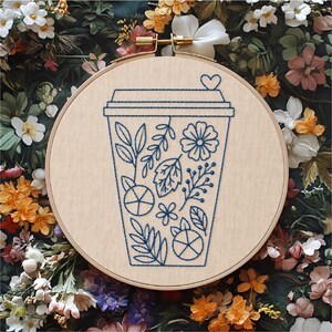 May include: A circular wooden embroidery hoop containing a light beige fabric panel. The fabric features a dark blue thread outline of a disposable coffee cup decorated with various botanical elements, including leaves, flowers, and berries.