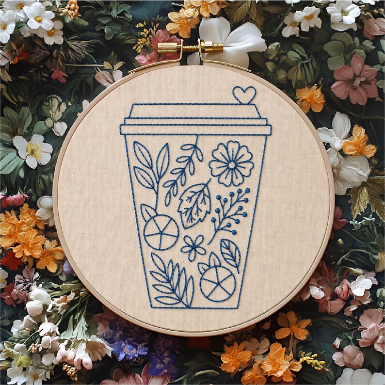 May include: A circular wooden embroidery hoop containing a light beige fabric panel. The fabric features a dark blue thread outline of a disposable coffee cup decorated with various botanical elements, including leaves, flowers, and berries.