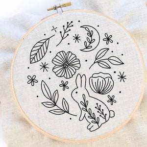 May include: A black and white embroidery hoop with a design of a rabbit, flowers, leaves, and a crescent moon. The design is stitched on a white fabric.