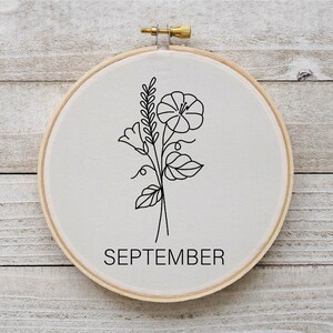 Puede incluir: Un aro de bordado blanco con un dibujo de l&iacute;nea negra de una flor y hojas. El texto "SEPTEMBER" est&aacute; impreso debajo del dibujo.