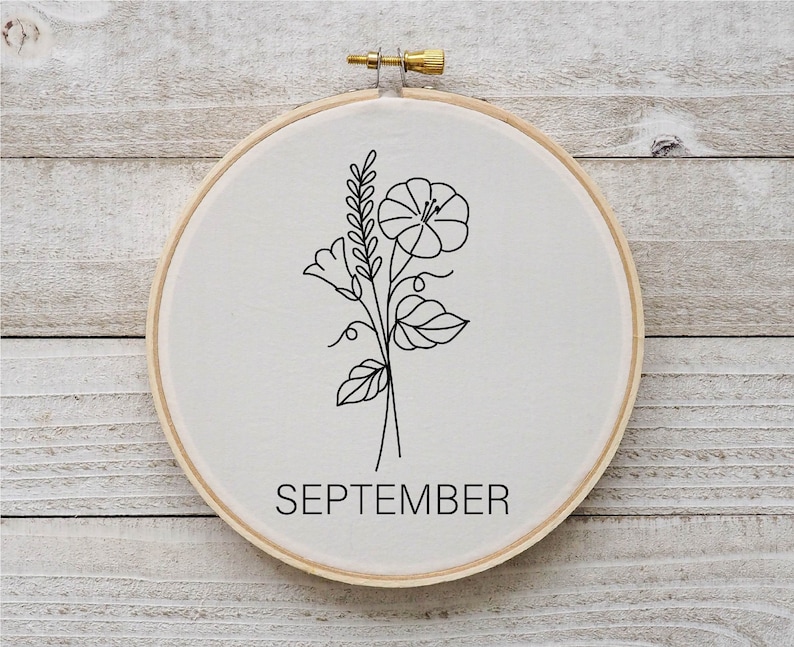 Puede incluir: Un aro de bordado blanco con un dibujo de l&iacute;nea negra de una flor y hojas. El texto "SEPTEMBER" est&aacute; impreso debajo del dibujo.