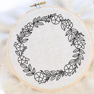 Könnte beinhalten: Ein schwarzer und weißer Stickrahmen mit einem floralen Kranzdesign. Der Kranz besteht aus Blättern und kleinen Kreisen.