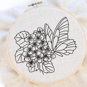 Op de afbeelding: Een ronde houten borduurring met een stuk witte stof. De stof is voorzien van een zwart lijnontwerp van een trosje kleine bloemen met bladeren en een grote vlinder met gedetailleerde vleugelpatronen aan de rechterkant.