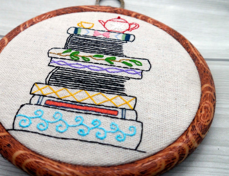 May include: Embroidered hoop art featuring a stack of books with a teapot and teacup on top. The books are decorated with colorful embroidery details, including flowers, leaves, and geometric patterns.