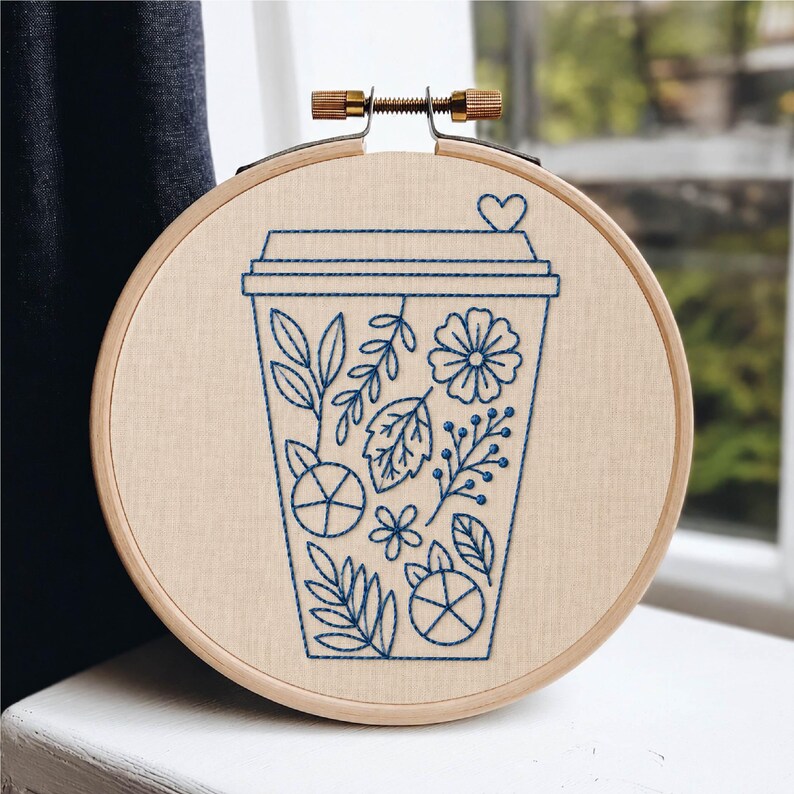 May include: A circular wooden embroidery hoop containing a piece of light beige fabric. The fabric features a dark blue thread outline of a disposable coffee cup decorated with various botanical elements including leaves, flowers, and a small heart.