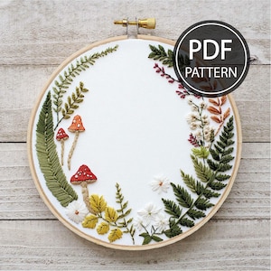 May include: An embroidered hoop art piece featuring a woodland theme. The design includes red-capped mushrooms, green ferns, and various flowers and leaves. The text "PDF PATTERN" is displayed on the upper right. The embroidery is set in a wooden hoop.