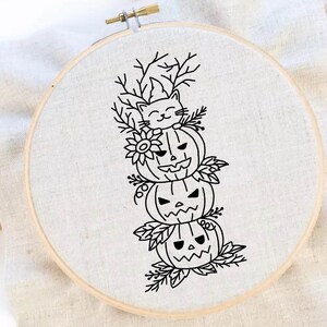 Könnte beinhalten: Ein schwarz-weißer Stickrahmen mit einem Halloween-Design. Das Design zeigt drei geschnitzte Kürbisse, die übereinander gestapelt sind und von einer lächelnden Katze gekrönt werden. Die Kürbisse sind mit Blättern und Blumen verziert.
