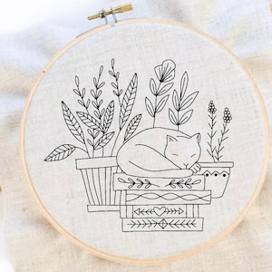 May include: Black and white embroidery hoop with a sleeping cat on a stack of books. The cat is surrounded by potted plants.
