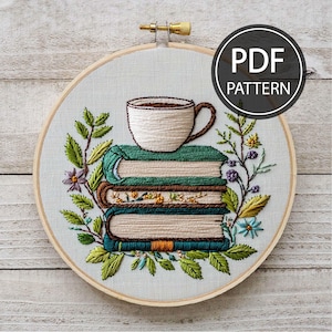 Puede incluir: Arte de bordado en aro que presenta una pila de libros con una taza de café en la parte superior, rodeada de bordado floral verde y morado. El aro está hecho de madera y tiene un fondo blanco.