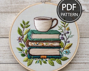 May include: Embroidered hoop art featuring a stack of books with a cup of coffee on top, surrounded by green and purple floral embroidery. The hoop is made of wood and has a white background.