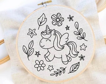 May include: A circular wooden embroidery hoop containing white fabric with a black line art design. The design features a central winged unicorn surrounded by various botanical elements including flowers, leaves, and small stars.