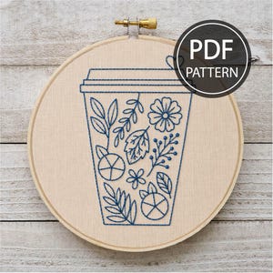 May include: A circular wooden embroidery hoop containing beige fabric with a blue thread outline of a coffee cup. The cup design is filled with various botanical elements including leaves, flowers, and berries. The text PDF PATTERN is visible.