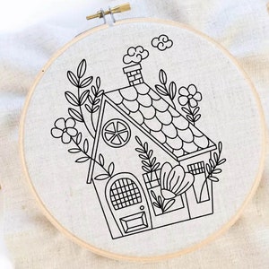 Könnte beinhalten: Ein schwarzer und weißer Stickrahmen mit einem Design eines kleinen Hauses mit einem Schornstein, einem Fenster in Form einer Zitrone und einer Tür mit einem Gittermuster. Das Haus ist von Blumen und Blättern umgeben.