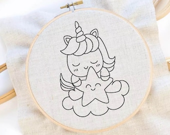 May include: A round wooden embroidery hoop containing a piece of white fabric with a black line art design of a unicorn with a horn, closed eyes, and a smiling mouth, sitting on a cloud and holding a smiling star with five points.