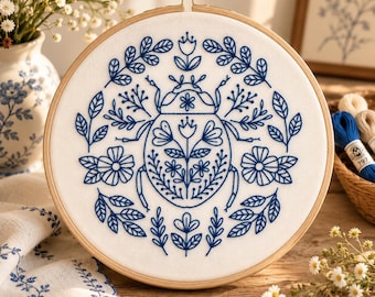 May include: A circular wooden embroidery hoop containing white fabric with a blue thread design of a beetle surrounded by symmetrical floral and leaf patterns. The beetle features intricate botanical details within its shell outline.