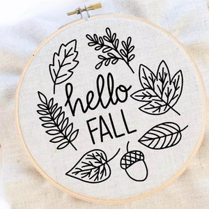 May include: A round embroidery hoop with a linen fabric featuring a black outline design of leaves and an acorn. The words "hello FALL" are written in a bold, handwritten style.