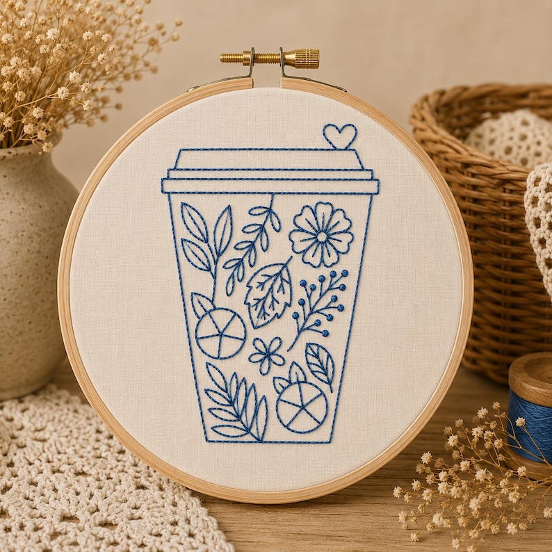 May include: A circular wooden embroidery hoop containing white fabric with a blue thread outline of a coffee cup. The cup design is filled with various botanical patterns including leaves, flowers, and berries, with a small heart on the lid.