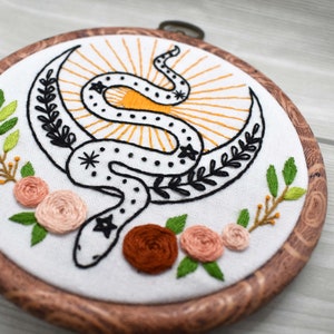 May include: Embroidered hoop art featuring a black snake wrapped around a crescent moon with a yellow sunburst behind it. The hoop is made of wood and has a natural finish. There are green leaves and pink flowers around the snake.