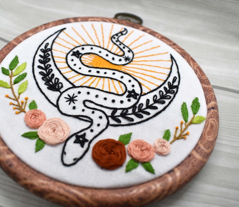 May include: Embroidered hoop art featuring a black snake wrapped around a crescent moon with a yellow sunburst behind it. The hoop is made of wood and has a natural finish. There are green leaves and pink flowers around the snake.