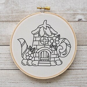 May include: Black and white line drawing of a teapot shaped house with a door and windows, surrounded by flowers. The drawing is on a white background and is framed in a wooden hoop.