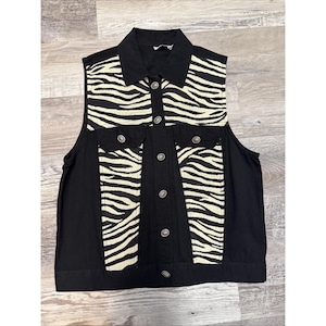 May include: A black denim vest with a zebra print design on the front panels and pockets. The vest has a collared neckline, silver buttons, and two front pockets. The vest is sleeveless and has a classic, casual style.