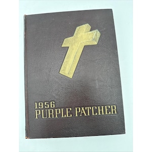 May include: A vintage book with a dark brown cover and a gold cross emblem. The title "1956 PURPLE PATCHER" is embossed in gold lettering. The book has a textured surface and appears to be a yearbook or commemorative item.