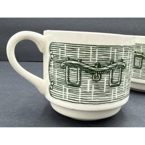 May include: Off-white ceramic mug with a green basket design. The mug has a curved handle and a stacked base. The basket design is detailed with horizontal and vertical lines, and a decorative handle.