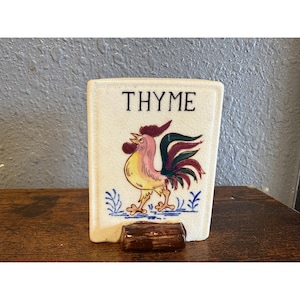 Może przedstawiać: Prostokątny ceramiczny słoik na przyprawy z napisem "THYME" i ilustracją koguta. Kogut jest żółty, różowy i czerwony, z niebieskimi akcentami. Słoik ma małą, brązową, prostokątną podstawę.