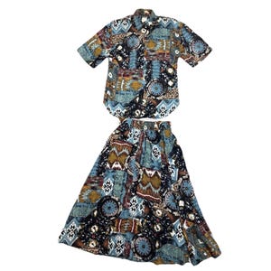 Pode incluir: Um conjunto de duas peças a condizer com uma camisa abotoada de manga curta e uma saia comprida. O conjunto tem um padrão geométrico colorido em tons de azul, castanho e bege. A saia é de estilo amplo e fluido.