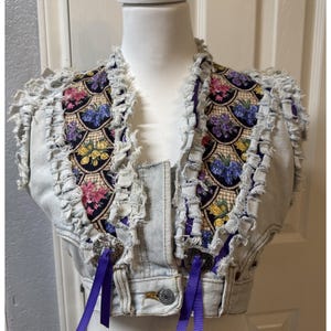 May include: Light blue denim vest with a floral patterned fabric on the collar and upper chest. The vest has a frayed edge design and purple ribbon accents. The floral pattern includes pink, yellow, and purple flowers.