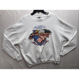 May include: White sweatshirt with a graphic featuring an American flag, an eagle, and the text "Proud to be American." The design incorporates red, white, and blue colors. The sweatshirt has long sleeves and a crew neck.