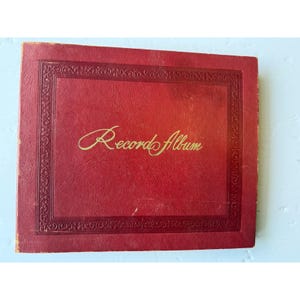 May include: A vintage red record album with gold lettering. The album cover has a decorative border and the words "Record Album" in a cursive font. The album appears to be made of textured material.