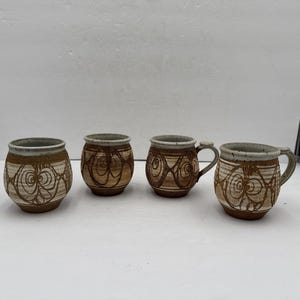 Könnte beinhalten: Vier Keramikbecher in Braun- und Cremetönen. Jeder Becher hat ein einzigartiges, handgezeichnetes Design aus wirbelnden Linien und Kreisen. Zwei Becher haben Henkel. Die Becher haben eine abgerundete Form.