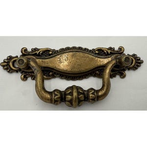 May include: Antique brass-toned drawer pull with ornate detailing. The pull features a curved handle and decorative backplate with scrollwork. The hardware is designed for furniture restoration or adding vintage charm to cabinets and drawers.