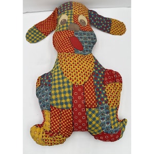May include: A patchwork dog-shaped pillow with a variety of colorful patterns. The dog has floppy ears, a red tongue, and brown eyes. The patchwork includes red, yellow, and blue fabrics with floral and star designs.