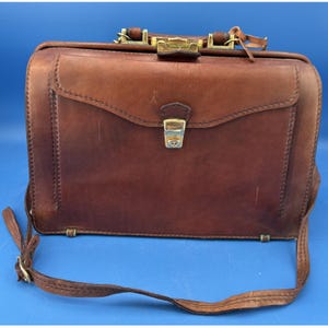 May include: A brown leather doctor-style bag featuring a structured rectangular frame, a top handle, and an adjustable shoulder strap. The bag includes a front flap with a gold-tone metal push-lock closure and visible stitching along the edges.
