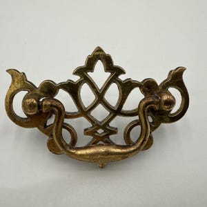 May include: Antique brass drawer pull with ornate filigree design. The pull features a curved handle and decorative flourishes on each side. The center has a diamond-shaped pattern. A vintage hardware piece.