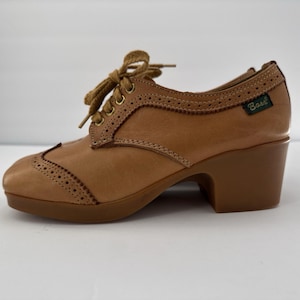 May include: A tan leather oxford shoe with a block heel, featuring brogue detailing along the toe cap and lace-up closure with tan laces. A small green tag with the word Bass is stitched onto the side of the shoe near the heel collar.
