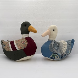 May include: Two duck-shaped doorstops. One has a green head, yellow beak, and burgundy body with floral accents. The other has a white head, pink beak, and blue body with lace trim. Decorative home decor.