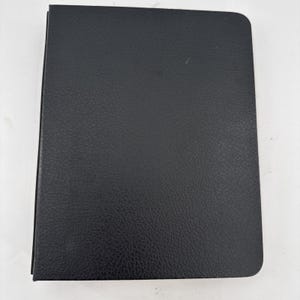 May include: A closed, black, leather-bound notebook with rounded corners. The cover has a textured surface. The notebook is rectangular and appears to be new.