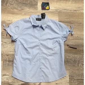 Op de afbeelding: Een lichtblauw overhemd met knopen, korte mouwen en strikdetails. Het overhemd heeft een klassieke kraag en een reeks knopen aan de voorkant. Een label is aan het overhemd bevestigd.