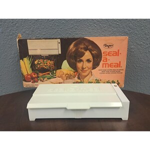 Peut inclure: Un appareil de scellage alimentaire blanc "Seal-a-Meal" avec le texte "seal-a-meal" sur le devant. La boîte derrière présente un portrait de femme et les mots "Dazey seal-a-meal". La boîte montre également des images d'aliments.
