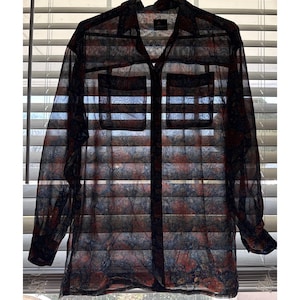 Puede incluir: Camisa transparente con botones y un patrón único. Presenta cuello, dos bolsillos en el pecho y mangas largas. El patrón combina tonos de rojo, azul y negro, creando un diseño visualmente interesante. La camisa es ligera y parece estar hecha de una tela delicada.